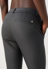 Personne portant un pantalon gris foncé ajusté avec des poches arrière et un petit logo métallique décoratif sur la poche droite, mains dans les poches avant.
