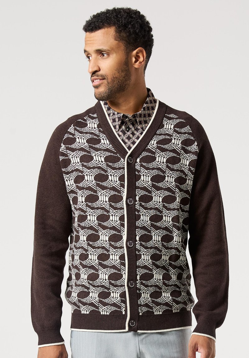 Hnědý kardigan s bílým geometrickým vzorem, s výstřihem do V, zapínáním na knoflíky a kontrastními okraji. Měkká pletená textura.