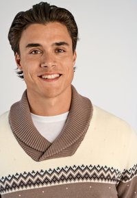 Gebreide trui met een crèmekleurig lichaam en brede bruine sjaalkraag. Beschikt over een geometrisch patroon in bruin en marineblauw over de borst.