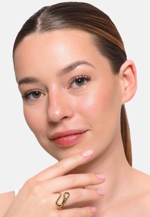 Junge Frau mit klarer Haut und glattem Haar berührt ihr Kinn und trägt einen goldenen Ring in abstrakter Form am Zeigefinger.