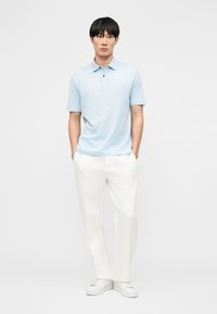 Egy férfi áll, kezeit zsebében tartja, világoskék pólóinget, fehér nadrágot és fehér tornacipőt visel, egyszínű fehér háttér előtt.