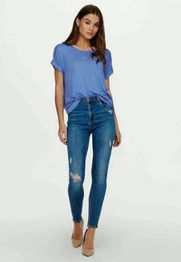 T-shirt azul de mangas curtas com um corte descontraído, combinado com jeans skinny azul escuro com detalhes rasgados. Sapatos de salto altos bege completam o look.