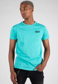 Türkises T-Shirt aus Baumwolle mit kurzen Ärmeln und Rundhalsausschnitt, mit einem schwarzen Logo auf der linken Brust. Getragen mit dunklen Jeans.