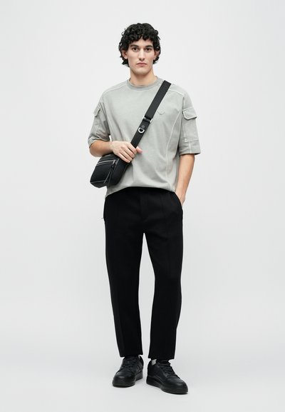 Γκρι κοντομάνικο πουκάμισο με μπαλώματα στους ώμους, μαύρο παντελόνι καλοραμμένο και μαύρα αθλητικά παπούτσια· το μοντέλο κρατάει μια μαύρη crossbody τσάντα με υφή.