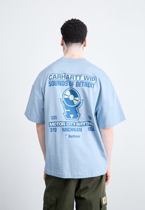 Mand iført en lyseblå Carhartt WIP skjorte med "Sounds of Detroit" og en vinylplade grafik på bagsiden, kombineret med oliven cargo bukser.