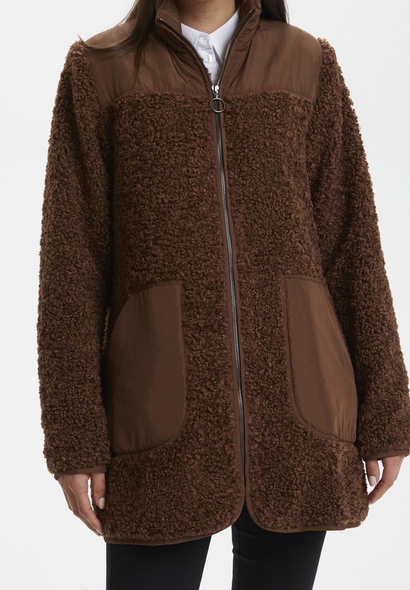 Veste polaire marron avec des patchs matelassés sur les épaules et les poches, fermeture éclair à l'avant, portée sur une chemise blanche à col.