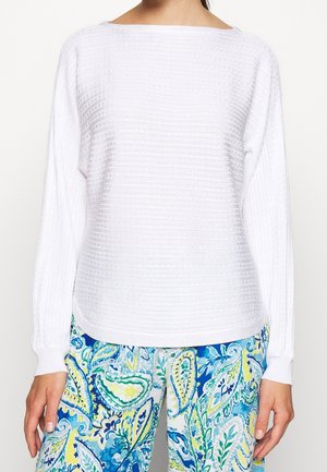 Persona con una camiseta blanca texturizada de manga larga y pantalones coloridos con estampado paisley en azul, verde y amarillo, recortados en el cuello y la cintura.