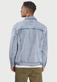 Chaqueta de mezclilla en azul claro con un corte clásico, costuras visibles y detalles de botones en los laterales. Presenta un cuello de pico y un detalle de parche en la parte posterior.