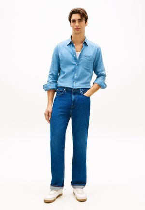 Lyseblå denimskjorte med en brystlomme, kombineret med lige, mørkeblå jeans. Hvide sneakers med lysebrune såler fuldender outfittet.