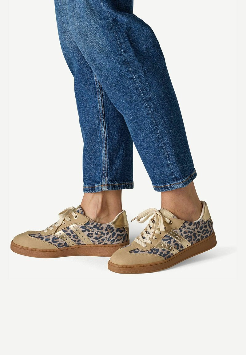 Personne portant un jean bleu et des baskets beiges avec des détails imprimé léopard, debout avec un pied légèrement levé.