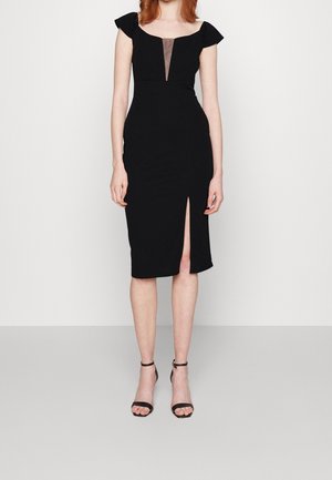 Woman wearing a fitted black dress with cap sleeves, a deep V mesh neckline, knee-length slit, and black ankle-strap heels.