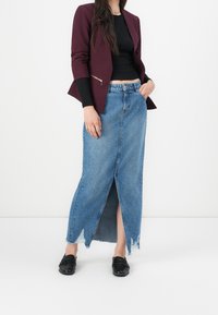 Jupe en denim avec une fente haute et un ourlet effiloché, associée à un haut noir ajusté et un blazer bordeaux avec une poche zippée. Mocassins noirs portés.
