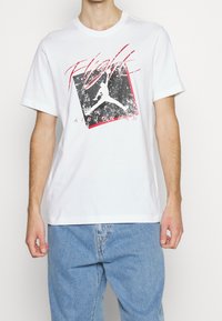 Camiseta de algodón blanca con mangas cortas, que presenta un diseño gráfico en negro y rojo del logo de Air Jordan y el texto "Flight".