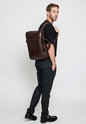 Mann trägt schwarzes Hemd, graue Hose und schwarze Schuhe, hält einen großen braunen Leder-Rucksack über der Schulter vor einfachem Hintergrund.