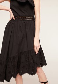 Frau in einem schwarzen ärmellosen Kleid mit Spitzen-Ausschneidungen an der Taille und am Saum, die mit der Hand auf der Hüfte vor einem hellen Hintergrund steht.