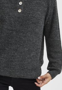 Dunkelgrauer Strickpullover mit zwei weißen Knöpfen auf der Brust, getragen von einer Person, die den Saum mit ihrer rechten Hand anpasst.