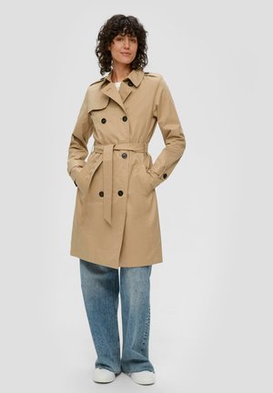 Trench-beige avec design à double boutonnage, taille ceinturée, poches latérales et boutons noirs. Assorti à un jean bleu ample et des baskets blanches.