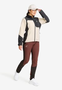 Woman in beige and black zip-up jacket, brown and black athletic pants, white sneakers, and light beige cap, walking with one hand adjusting her cap.