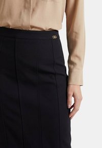 Gonna nera con righe sottili verticali e dettaglio del logo dorato sulla vita, abbinata a una camicetta beige a maniche lunghe.