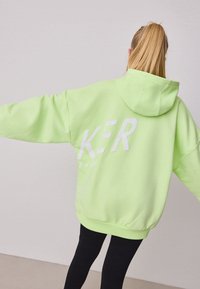 Neonzöld, oversized kapucnis pulóver fehér felirattal a hátoldalán. Húzózsinóros kapucnival és bordázott mandzsettákkal rendelkezik, puha polár anyagból készült.