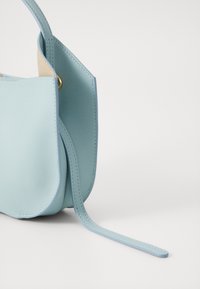 Sac à main en cuir bleu clair avec une poignée unique, des bords incurvés et une longue bandoulière s'étendant vers l'extérieur sur un fond blanc.