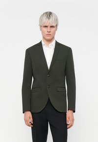 Mørkegrøn blazer med to knapper, reverskrave og to forlommer, parret med en hvid skjorte med krave og sorte bukser.