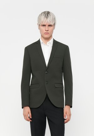 Blazer vert foncé à deux boutons, avec des revers en notch et deux poches avant, associé à une chemise blanche à col et un pantalon noir.