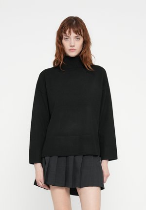 Svart turtleneck genser med ribbet søm og mansjetter, kombinert med en plisserte mørk grå skjørt. Myk tekstur; avslappet, oversized passform.