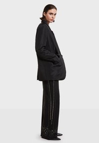 Crni dugi kaput opuštenog kroja s velikim džepovima, uparen s crnim hlačama koje imaju metalnu vezicu i šiljaste nosove.