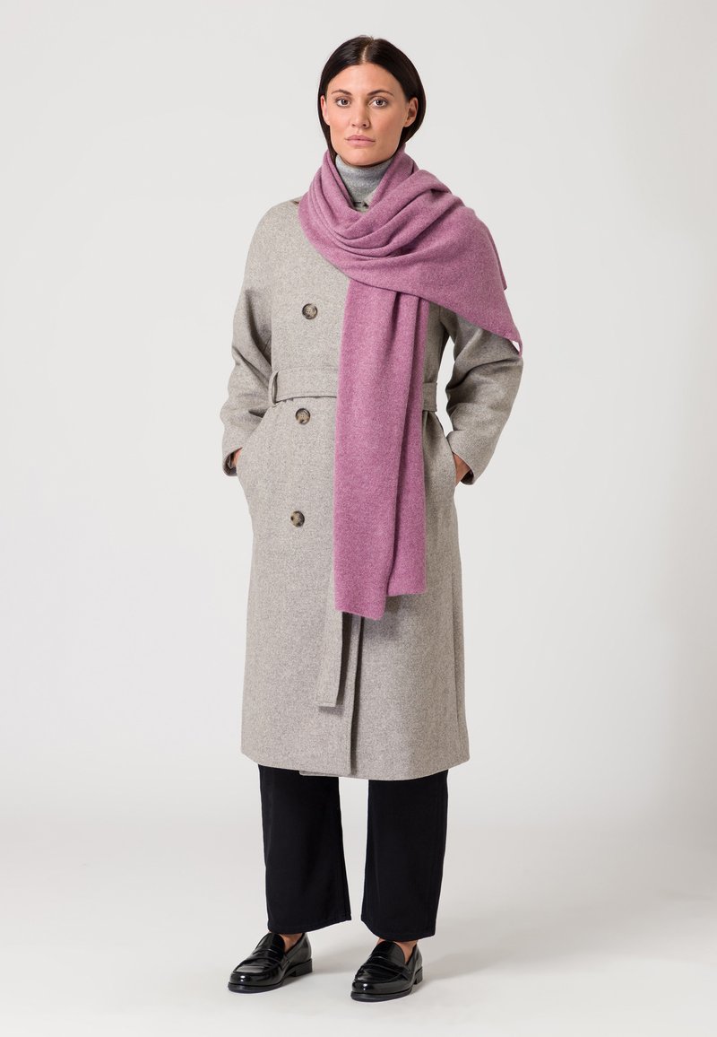 Cappotto grigio in lana con bottoni doppi, vita svasata e cinture, abbinato a una morbida sciarpa rosa a trama. Pantaloni neri e mocassini completano il look.