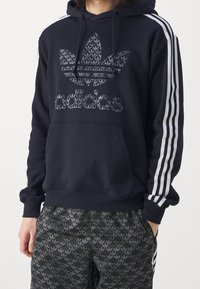 Navy hoodie with a textured black Adidas logo and three white stripes on the sleeves. Features a kangaroo pocket and ribbed cuffs.