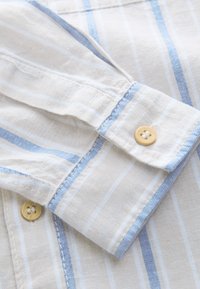 Približan posnetek manšete bele srajce z modro-belimi navpičnimi črtami in bež gumbi, ki prikazuje teksturo tkanine in detajle šivanja.