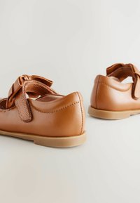 Beige Leder Schuhe mit glatter Textur. Verfügt über eine Schleifenakzent und einen verstellbaren Riemen, mit einer hellbraunen Sohle und runder Form.