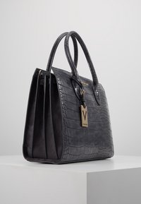 Svart krokodilprickat läderhandtag med två topphandtag, en texturerad yta och en guldfärgad logotagg fäst framtill.