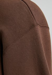Bruin sweatshirt stof met een close-up van een schoudernaad, met een zachte, gestructureerde afwerking en nette stikdetails.