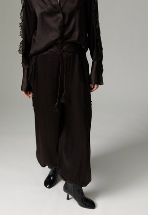 Personne portant un pantalon ample en satin noir avec une taille à cordon, un haut à manches longues assorti avec bordure en dentelle, et des bottines noires à talons.