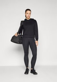 Svart hettegenser med snøring, kombinert med matchende leggings og svarte treningssko. Modellen holder en svart treningsbag, plassert mot en grå bakgrunn.
