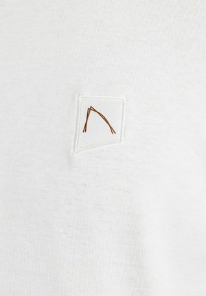 Valkoinen puuvillapaita, jossa on pieni, neliömäinen pala, jossa on ruskealla ommeltu geometrinen kuvio. Silkkinen rakenne, jossa on minimimäärä yksityiskohtia.