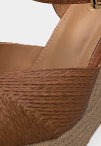 Sandale compensée tressée marron avec bride à la cheville et semelle intérieure beige rembourrée, présentant un détail tressé texturé sur la semelle plateforme.