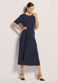 Navy blauwe midi-jurk met korte mouwen, geknoopte detail bij de taille en een gladde, soepele stof. Gecombineerd met zwarte hoge hakken.