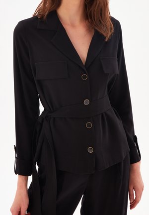 Woman wearing a black belted shirt jacket with gold buttons and flap pockets, paired with matching black pants against a white background.
