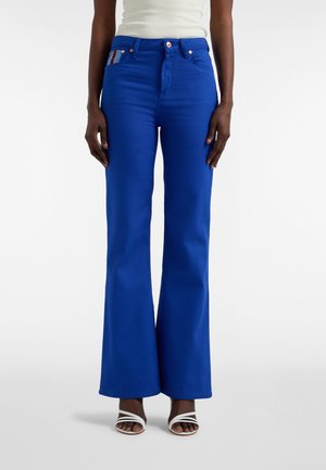 Dzwony niebieskie jeansy wykonane z wytrzymałej tkaniny, z wysokim stanem, ozdobione kieszenią w paski oraz kontrastowym guzikiem. Połączone z białymi sandałami.