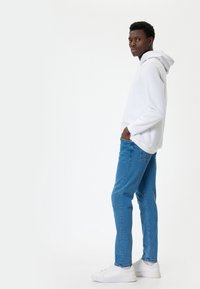 Sudadera blanca con capucha, vaqueros azules y zapatillas blancas. El conjunto presenta un diseño simple y un corte holgado, con una textura suave.