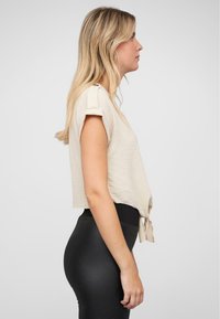 Cropped top beige corto con maniche arrotolate, caratterizzato da un dettaglio a nodo sull'orlo, abbinato a leggings neri a vita alta.
