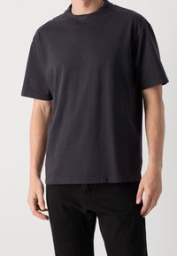 Homme portant un t-shirt noir uni à manches courtes avec le logo "BOSS" sur le col et un pantalon noir, debout devant un fond uni.