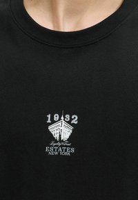 Sorte bomulds t-shirt med rund hals; har hvid grafisk tekst og et bygningsdesign, inklusive året "1932" og "ESTATES NEW YORK."