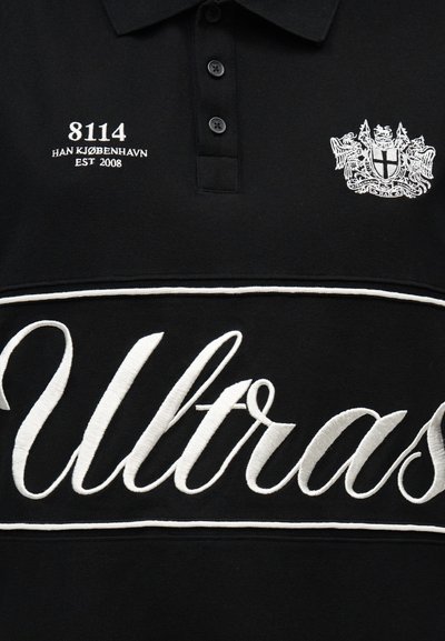 Μαύρο πόλο πουκάμισο με λευκή κεντημένη επιγραφή "8114 HAN KJØBENHAVN EST 2008," ένα έμβλημα με κονκάρδα και μεγάλο γράψιμο "Ultras" μπροστά.