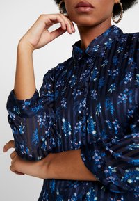 Námořnická květinová blůza s vertikálními pruhy, zdobená modrými a růžovými květy. Zapínání na knoflíky, špičatý límec a nařasené rukávy.