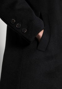 Melns vilnas mētelis, kurā redzama tuvplānojuma detaļa ar piedurkni, kas rotāta ar trim melniem pogām un sānu kabatu, izceļot mīkstu tekstūru un pielāgotu dizainu.