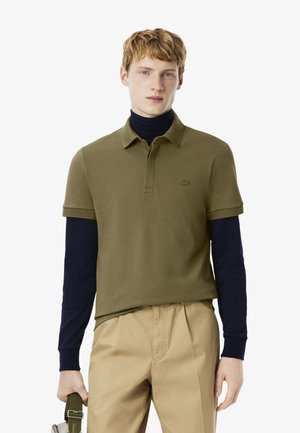 Junger Mann mit kurzen roten Haaren, der ein olivgrünes Poloshirt über einem schwarzen Rollkragenpullover und beigen Hosen trägt, mit einer Taschenriemen in der linken Hand.
